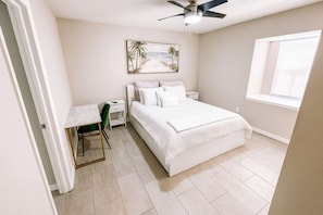 Ein Schlafzimmer mit Bett, Schreibtisch, Stuhl, Nachttisch, Deckenventilator und Fenster.