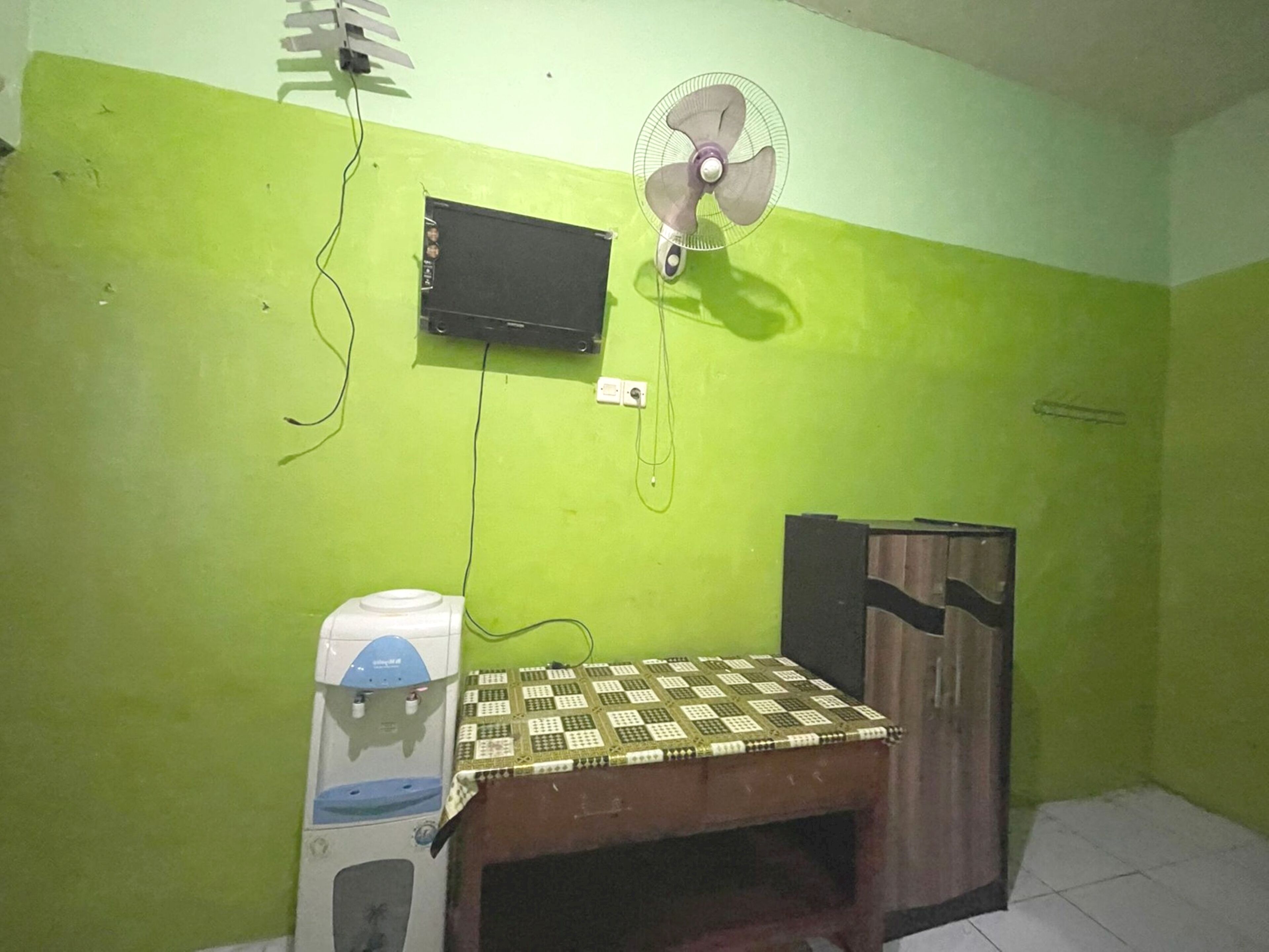 A room with a green wall, a bed with a patterned bedspread, a white water dispenser, a black TV mounted on the wall, and a standing fan.