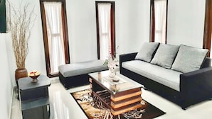 Ein modernes Wohnzimmer mit grauem Sofa, einem Couchtisch und einem Bücherregal. Drei Fenster mit Vorhängen, eine Vase mit Blumen und eine hohe Vase mit Zweigen sind ebenfalls zu sehen.