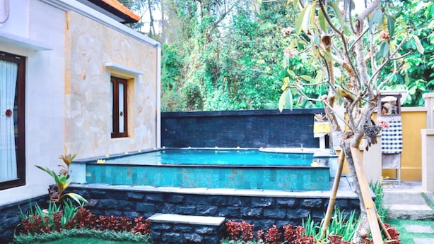 Ein Schwimmbad mit Steinmauer und Holzdeck, umgeben von einem Garten mit roten Blüten und einem Baum.