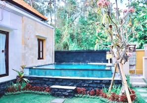 Une piscine bordée d’un mur en pierre et dotée d’une terrasse en bois, le tout entouré d’un jardin agrémenté de fleurs rouges et d’un arbre.