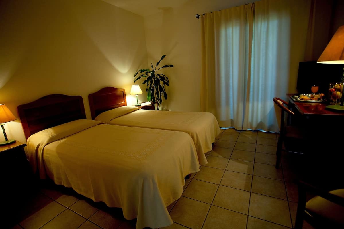 A hotel room with two beds, a desk, a chair, a lamp, and a potted plant.