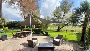 Ein Terrassenbereich im Freien mit Tisch und Stühlen, einem großen Sonnenschirm und einem grünen Rasen im Hintergrund.