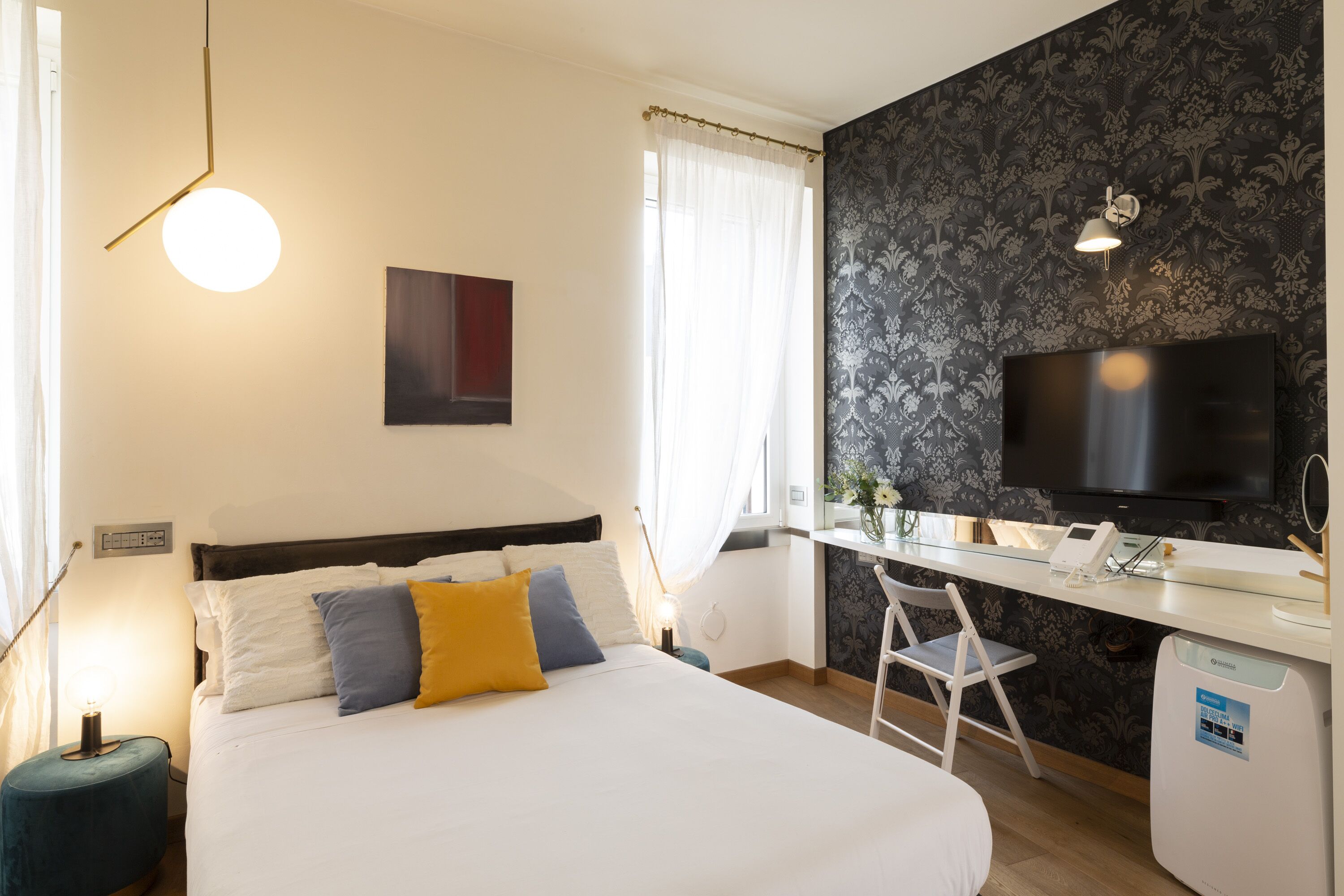 A hotel room with a bed, a desk with a flat-screen TV, a chair, a small table, and a wall-mounted TV.