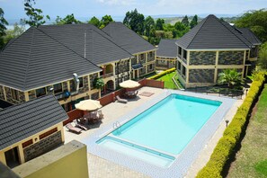 A large swimming pool with a tiled deck, surrounded by lounge chairs and umbrellas, and adjacent to a building with a dark roof. - Epashikino Resort and Spa (Gilgil)