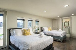 Two double beds with white bedding and decorative pillows, a small table, and a bathroom with a mirror and sink.