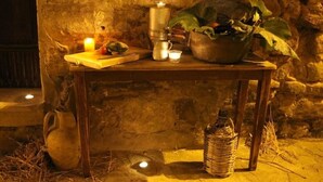 Een rustieke stenen muur met een houten tafel waarop een kaars, een metalen doos, een bloempot en een gevlochten mandje staan.