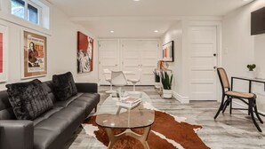 A modern living room with a leather sofa, glass coffee table, and a cowhide rug. There are white doors in the background, a TV mounted on the wall, and a framed poster on the wall. - Hilltop I Cherry Creek I Guest suite I Museums (Denver)
