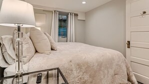 A bedroom with a large bed, a bedside table with a lamp, a glass table with a remote control, and a view of the outdoors through a window with curtains. - Hilltop I Cherry Creek I Guest suite I Museums (Denver)
