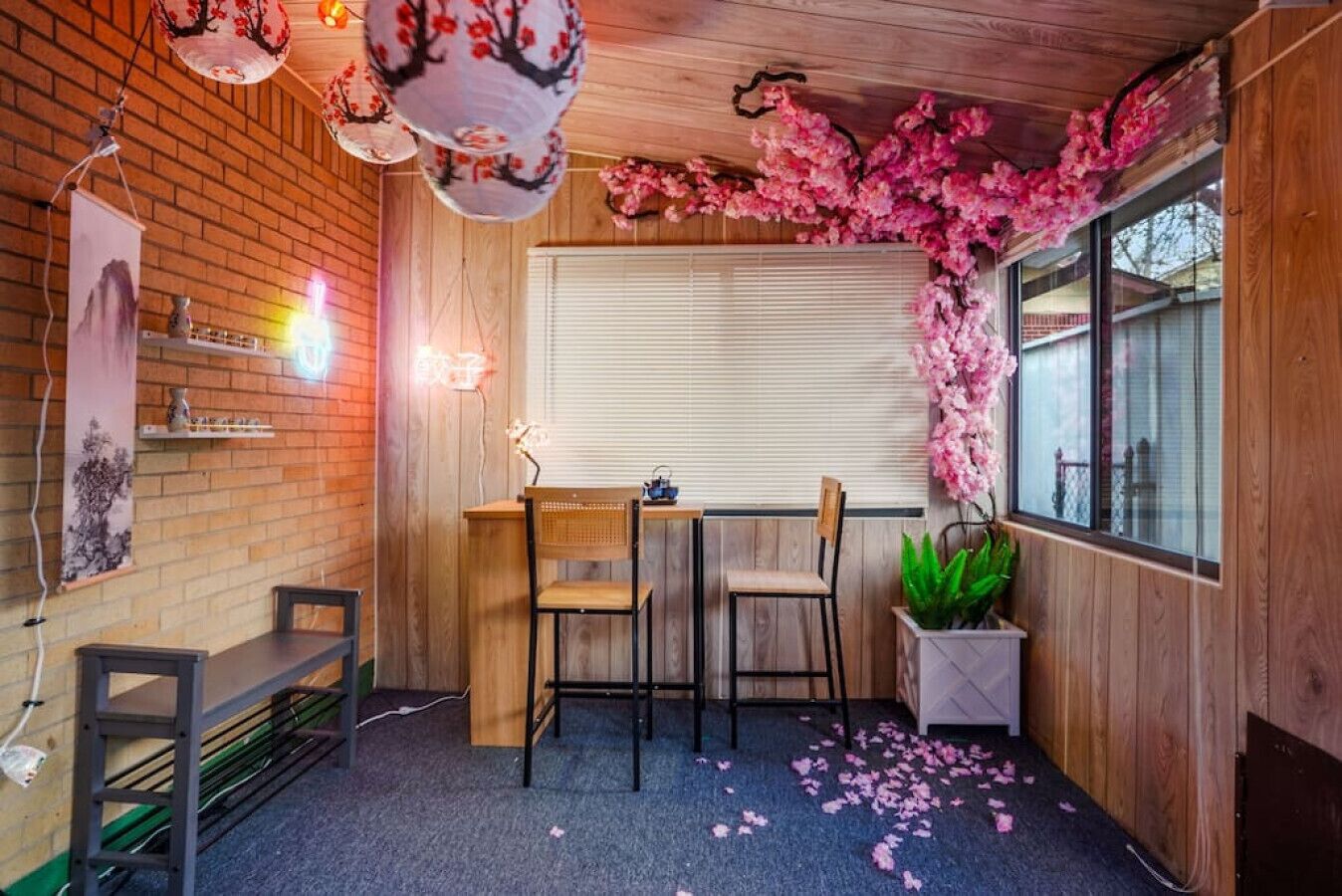 A cozy room with a wooden desk, two chairs, a window with blinds, and a decorative pink floral arrangement.