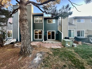 A two-story building with a large tree in the foreground, a paved patio, and a covered outdoor area with a heater. - Stylish Townhouse Retreat - 15 min to DT (Westminster)