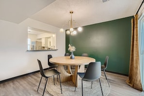 A dining area with a round table, six chairs, a vase with flowers, a hanging light fixture, and a mirror on the wall.