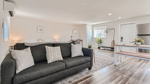 A modern living room with a dark grey sofa, a wooden coffee table, and a kitchen area with a stainless steel refrigerator.