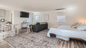 A modern, well-lit bedroom with a large bed, a grey sofa, a small table, and a TV mounted on the wall.