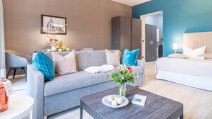 A modern hotel room with a grey sofa, a bed with a patterned headboard, a coffee table with a book and decorative items, and a painting on the wall. - Appartement Himmelblau mit Blick auf Bodden und Ostsee im Ostseebad Wustrow (Wustrow)