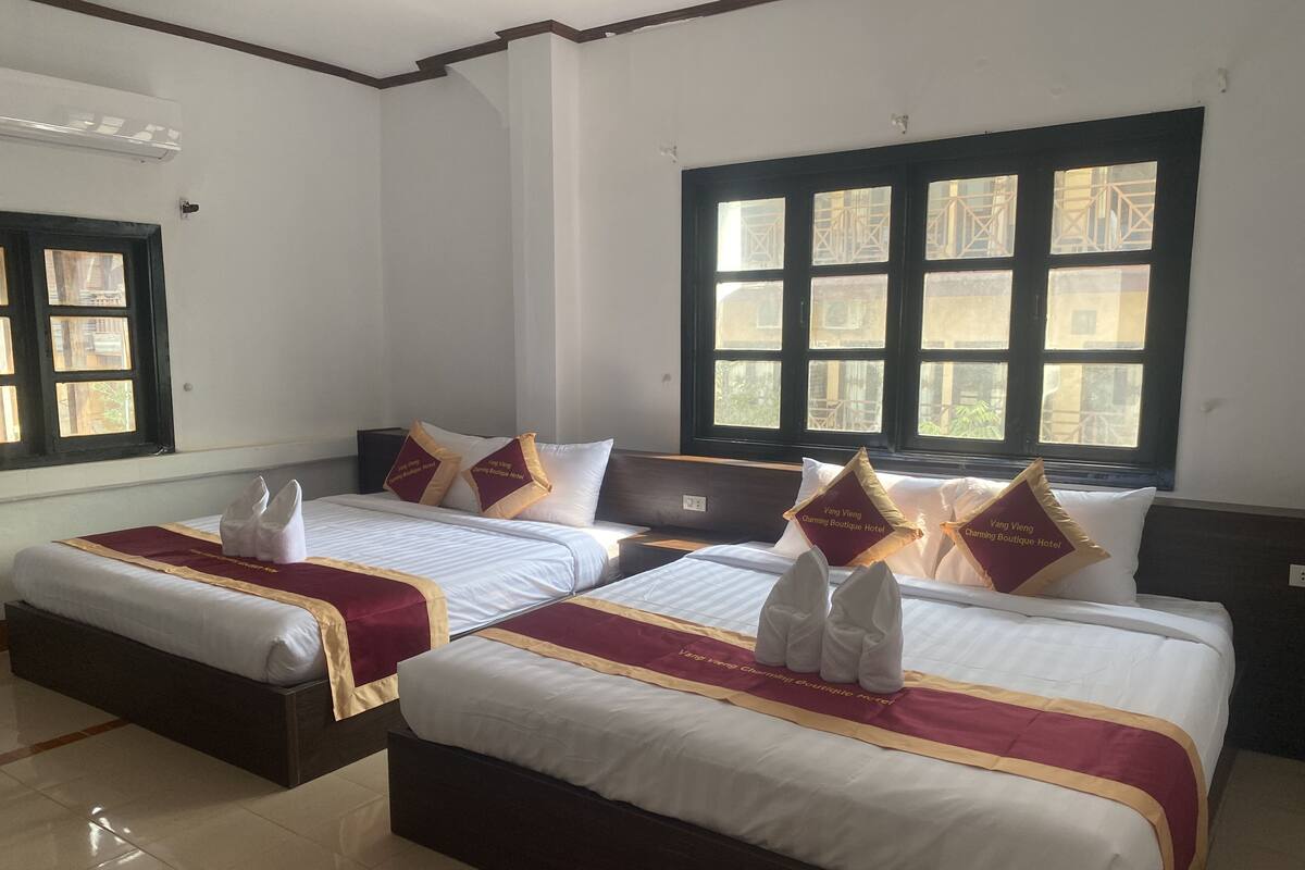 A hotel room with two beds, each with white linens and maroon bedspreads. There are decorative pillows with text, a window with a view of buildings, and a tiled floor.