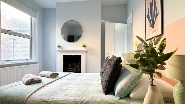 A bedroom with a bed, a fireplace, a round mirror, and a vase with flowers. - Spacious & stylish 3-bed home in Centre of Canterbury (Canterbury)