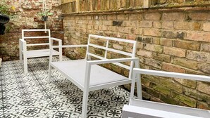 A patio area with two white metal chairs and a table, set against a brick wall with a wooden fence. - Spacious & stylish 3-bed home in Centre of Canterbury (Canterbury)