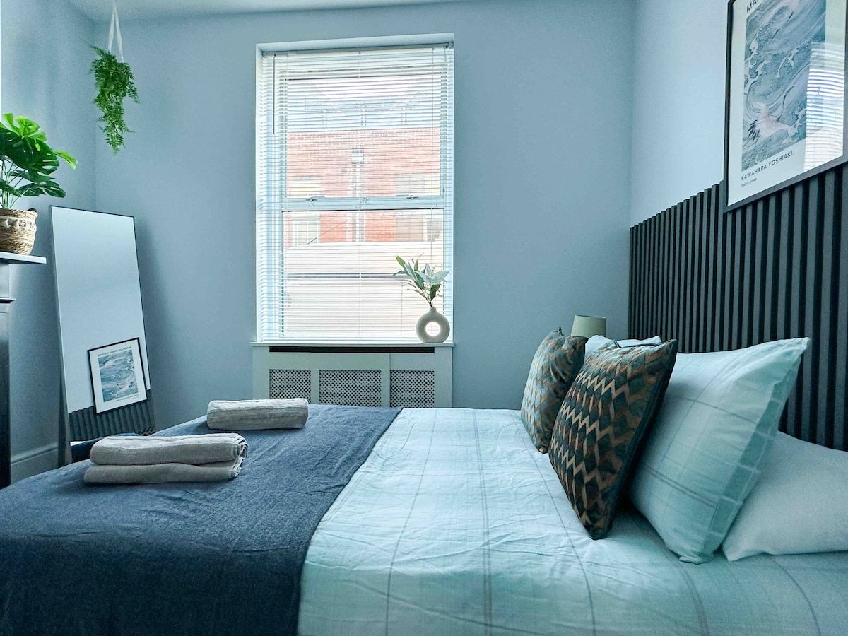 A bedroom with a bed, pillows, a bedside table, a lamp, a window, and a plant.
