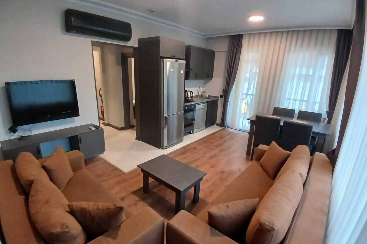 A living room with a sofa set, a television, a kitchen area, and a dining table with chairs.