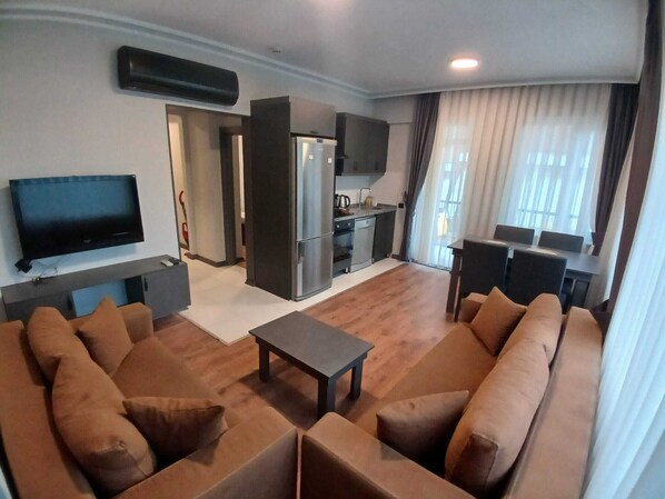 A living room with a sofa set, a television, a kitchen area, and a dining table with chairs.