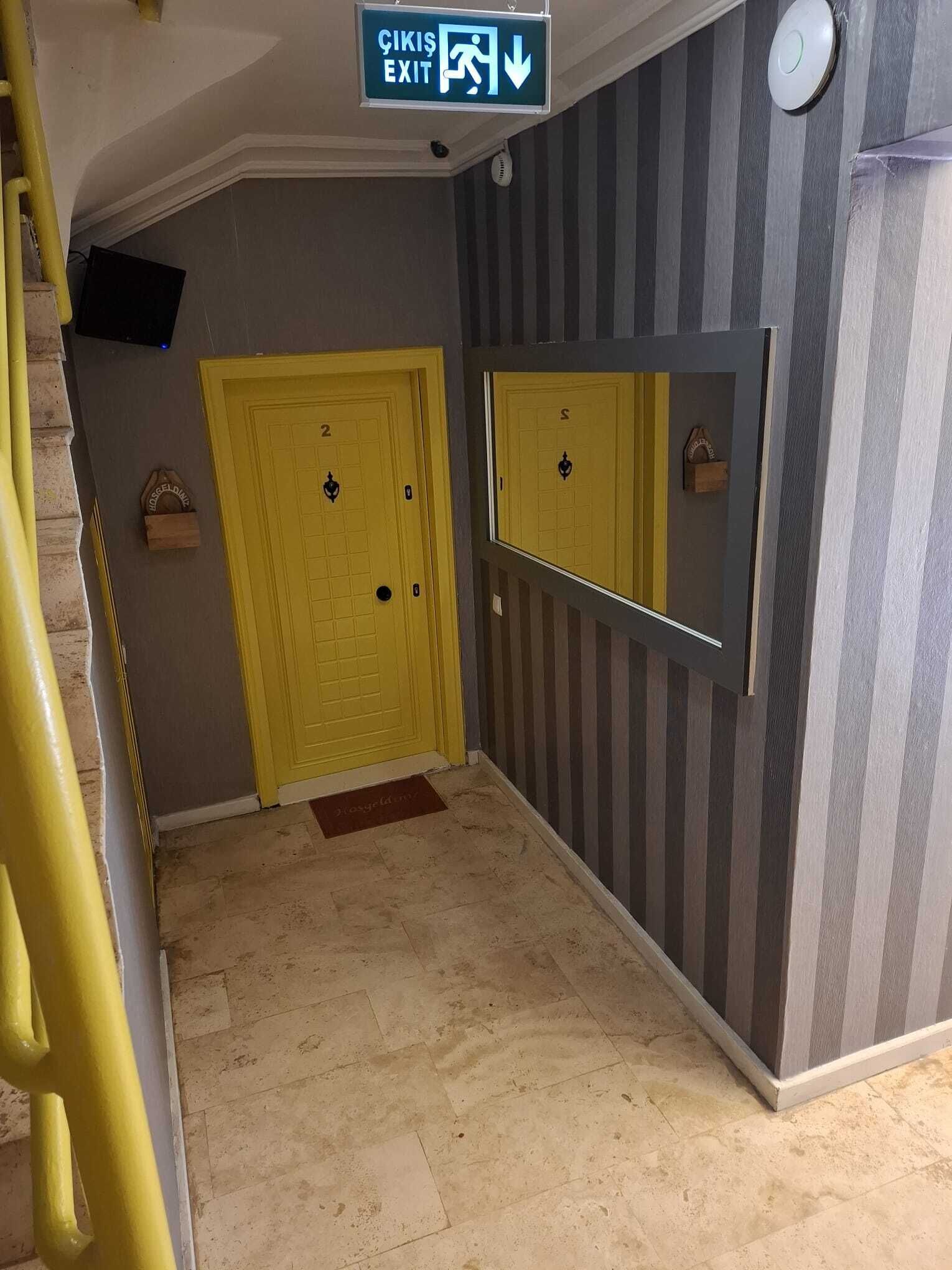 Un pasillo con una puerta amarilla marcada con el número 2, un muro espejado y un cartel que indica 'ÇIKIŞ EXIT'.