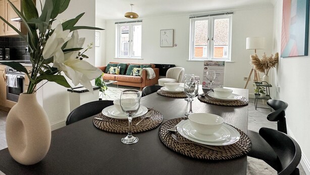 A dining area with a round table set for four, surrounded by woven placemats, with a vase of white flowers on the left.