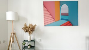 A room with a floor lamp, a vase with dried flowers, a small shelf with a plant, and a colorful abstract painting on the wall.
