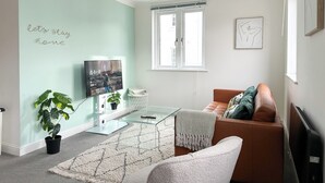 A living room with a teal wall, a glass coffee table, a brown leather sofa, a white armchair, and a large window.