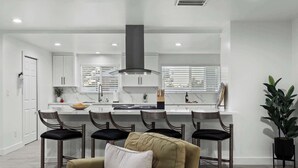 A modern kitchen with white cabinets, a central island with bar stools, and a dining area with a sofa and chairs. - Private Pickleball, B-Ball, Putt, Pool, Spa, Grill (Scottsdale)