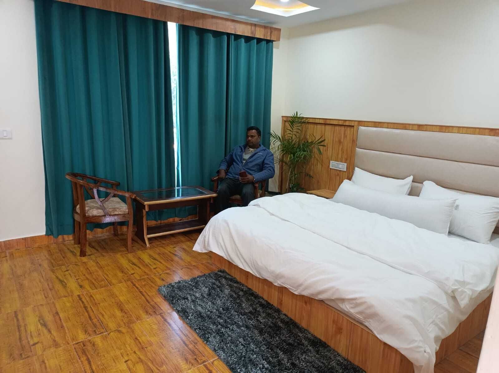 A hotel room with a bed, a wooden chair, a small table, and a large window with teal curtains.