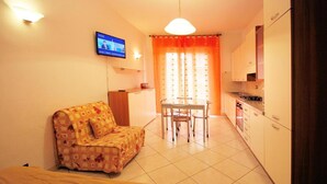 A compact living space with a sofa, a dining table with chairs, a TV mounted on the wall, and a kitchen area with cabinets and a stove. - Europa 29 Trilocale Manhattan (Loano)