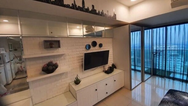 A modern living room with a flat-screen TV, white brick wall, and a balcony with a city skyline view. - Capital O 93636 La Grande Apartment By UJM (Bandung)