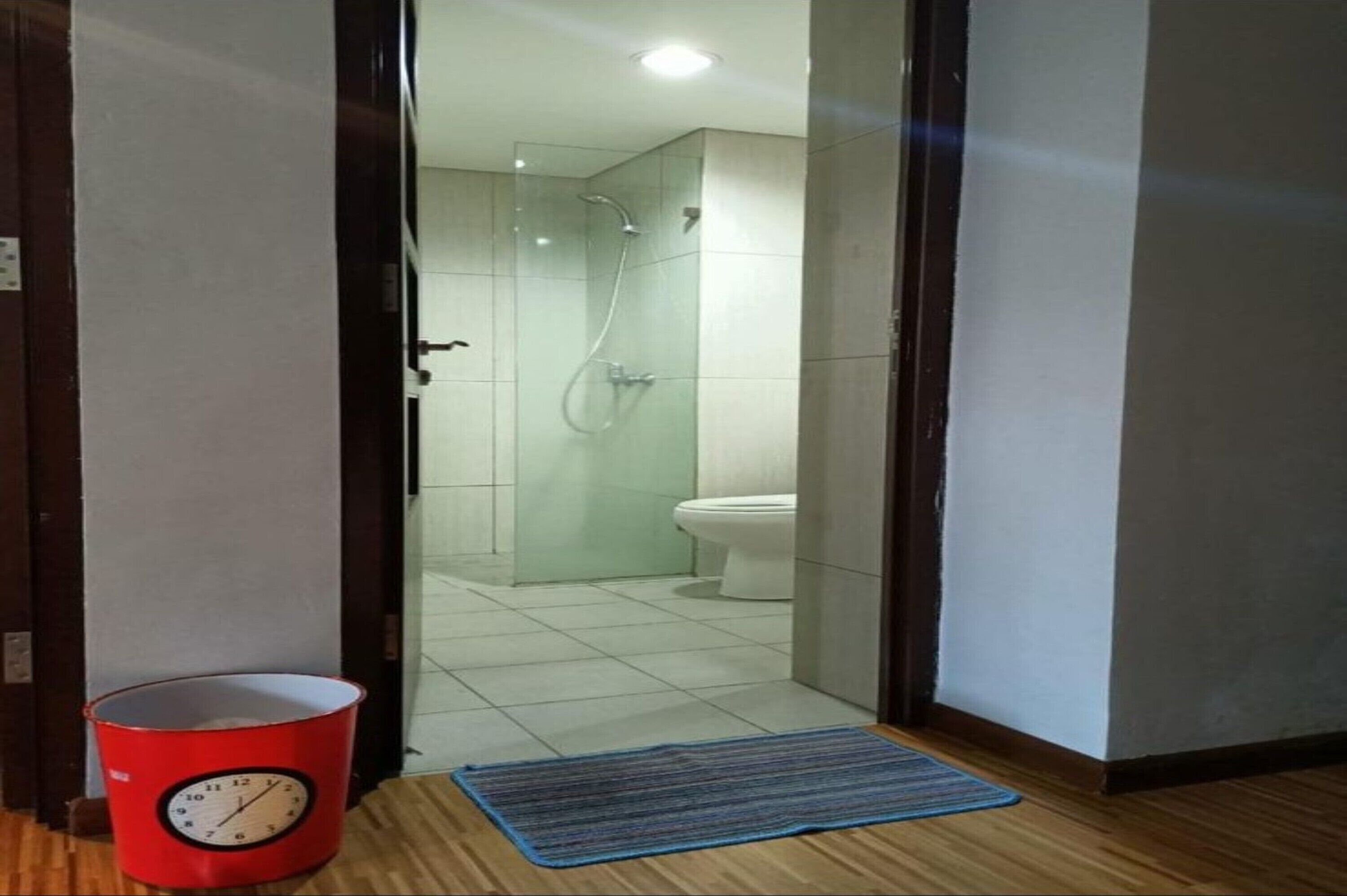 A bathroom with a glass shower enclosure, a white toilet, and a red bucket with a clock.