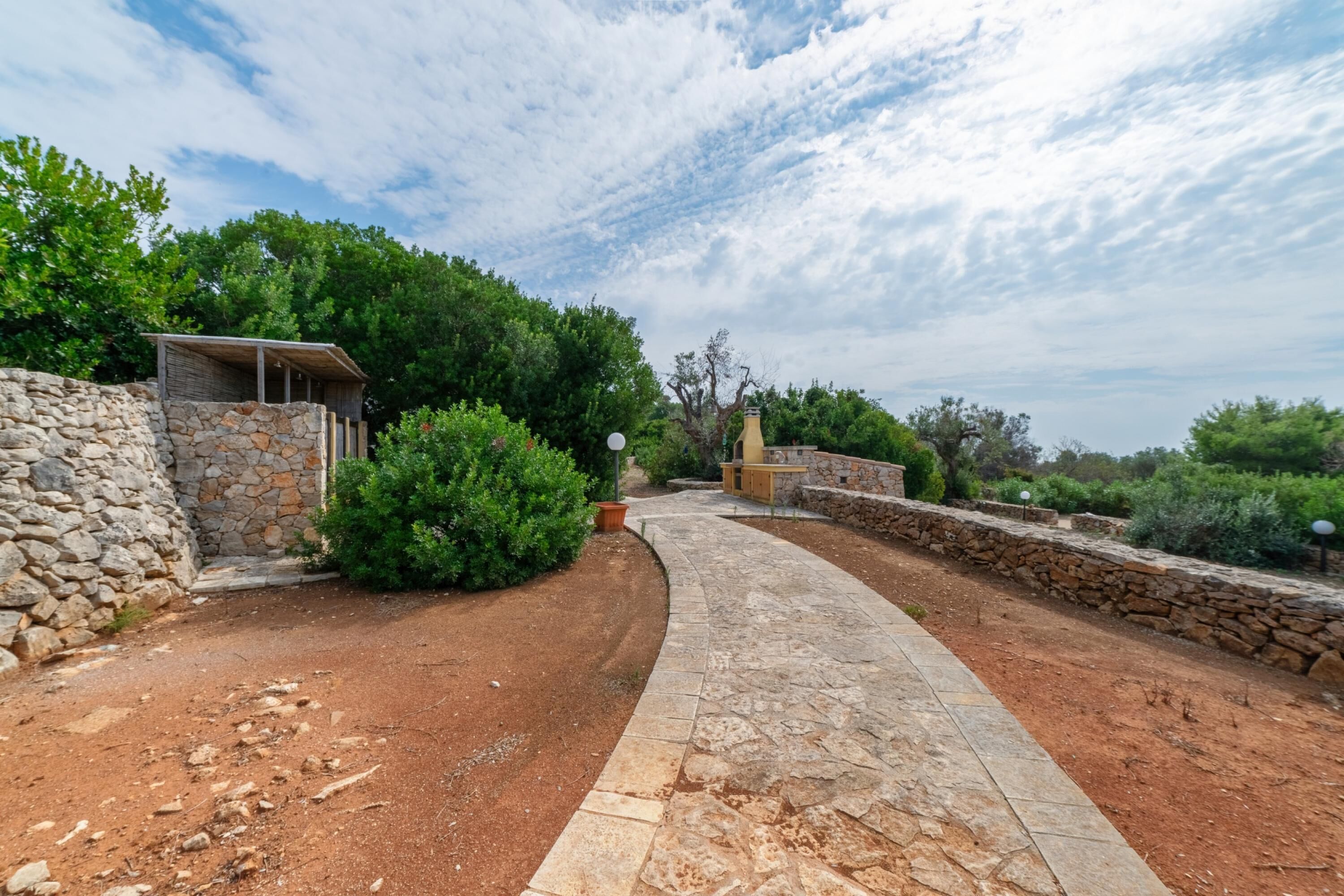 A stone pathway leading to a rustic building with a stone wall and greenery.