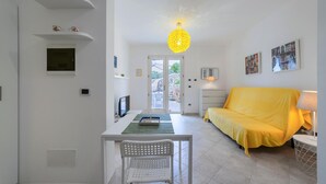 Ein Zimmer mit Esstisch, gelber Couch, weißem Stuhl, einer Pendelleuchte und einer zur Außenseite führenden Tür.