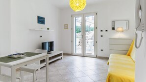 Ein kleiner Raum mit einem weißen Tisch, einem Fernseher, einem gelben Bett und einem großen Fenster mit Blick auf Grünflächen.