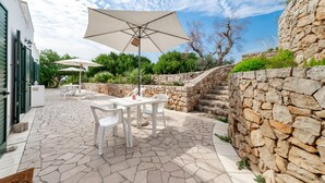 Eine Terrasse mit Steinboden, weißen Gartenmöbeln und einem großen Sonnenschirm.