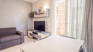 A modern living room with a sofa, TV, and dining table, featuring a stone wall and a sliding glass door. - Il Vento del Sud - Italian Homing (Padenghe sul Garda)