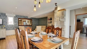 A dining area with a wooden table set for six, surrounded by wooden chairs. There is a stone accent wall, a kitchen in the background, and a mirror above the table. - Gîte Spacieux 3 Chambres, en Bord de Mer, Bretagne Sud, 56, Port-louis (Port-Louis)