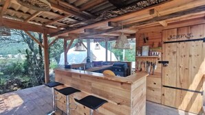 Ein rustikaler Barbereich aus Holz mit Grill, drei Hockern und Blick ins Freie.