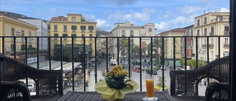 Ein Balkon mit Korbmöbeln, einem Tisch mit einer Blumenvase und einem Glas Orangensaft, und Blick auf eine Straße mit Gebäuden und einem Glockenturm.