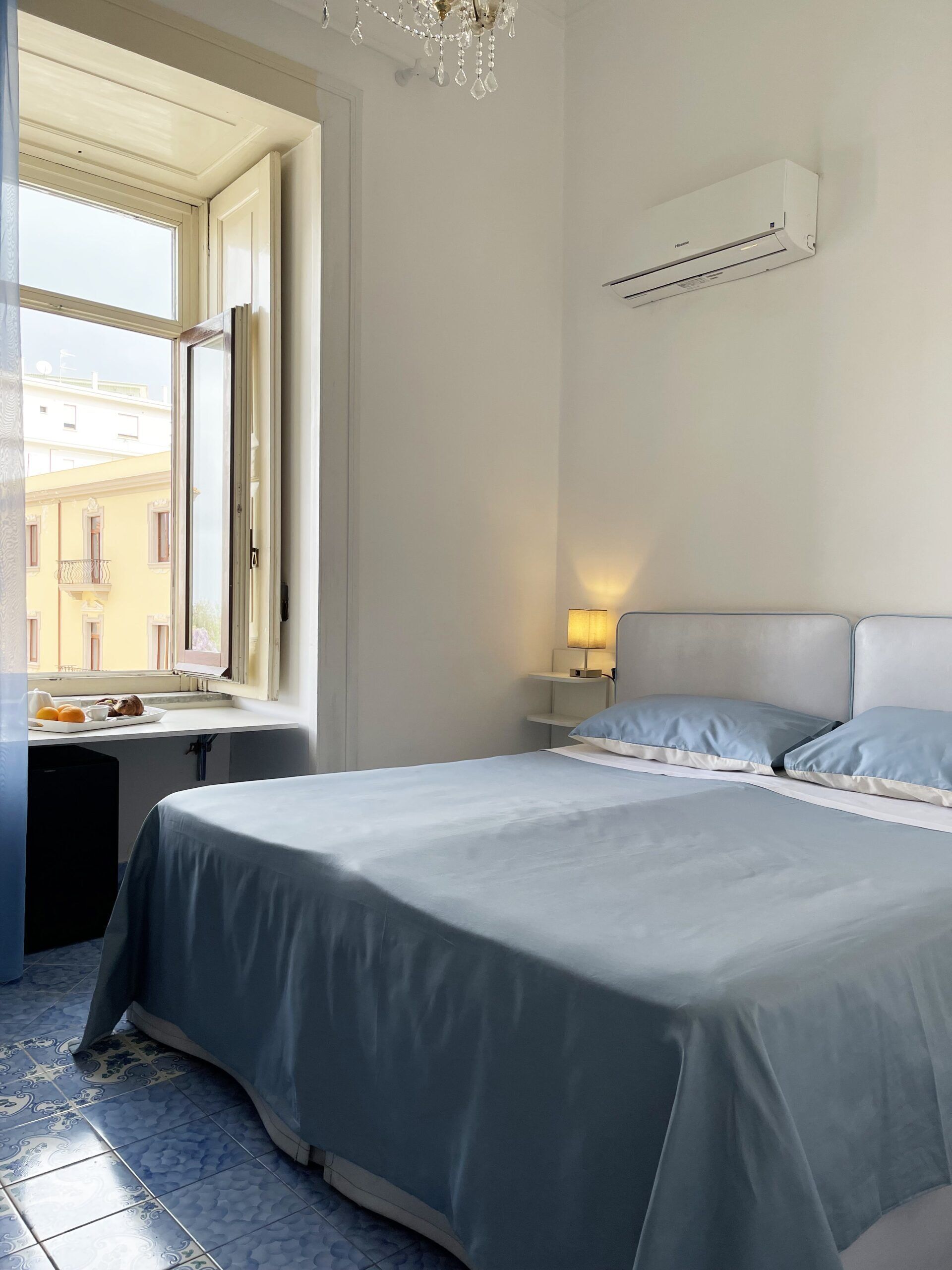A bedroom with a bed, a nightstand, a lamp, and a window with a view of buildings outside.