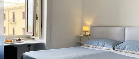 A bedroom with a bed, a nightstand, a lamp, and a window with a view of buildings outside.
