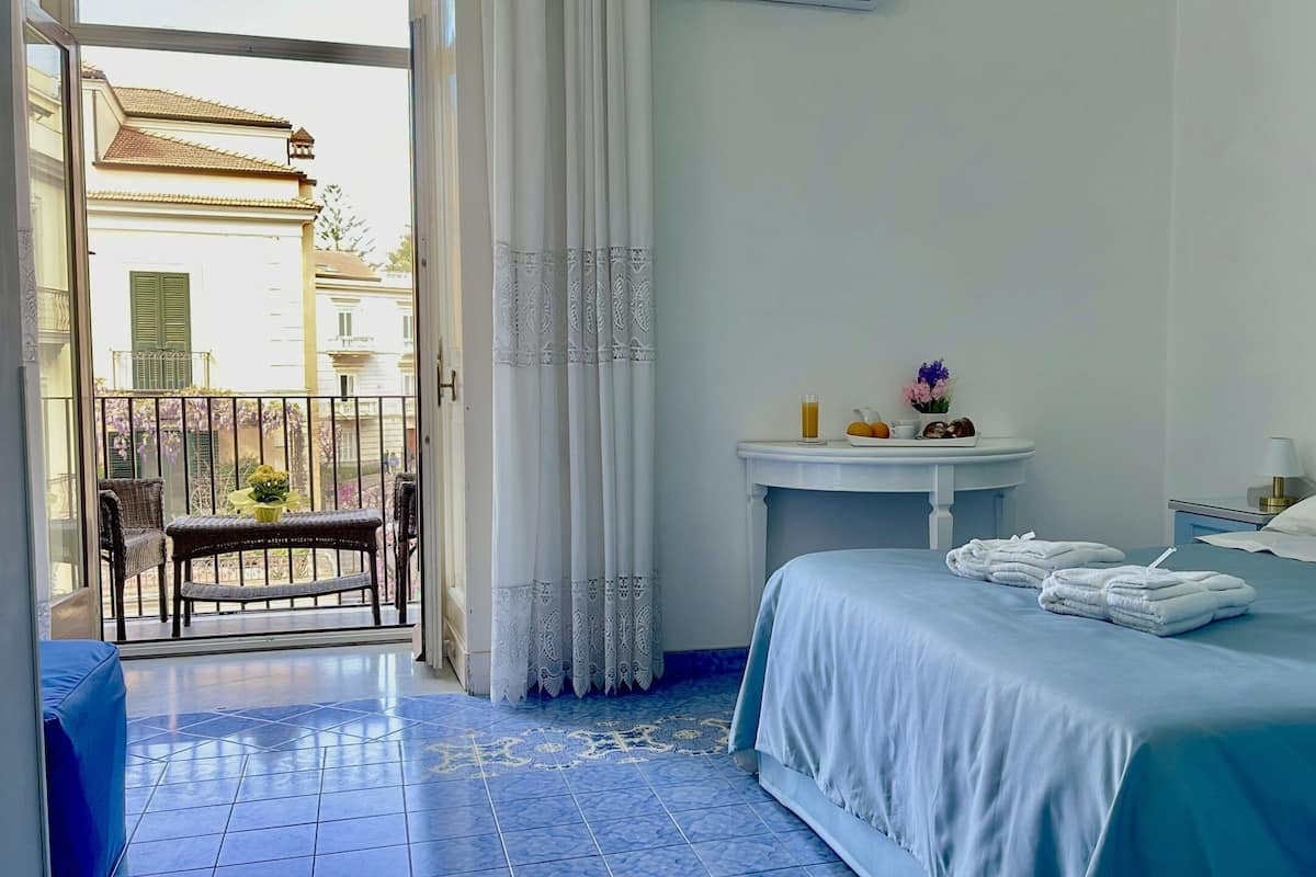 Habitación de hotel con cama, una mesita redonda con un florero, una silla y un balcón con vistas a edificios.