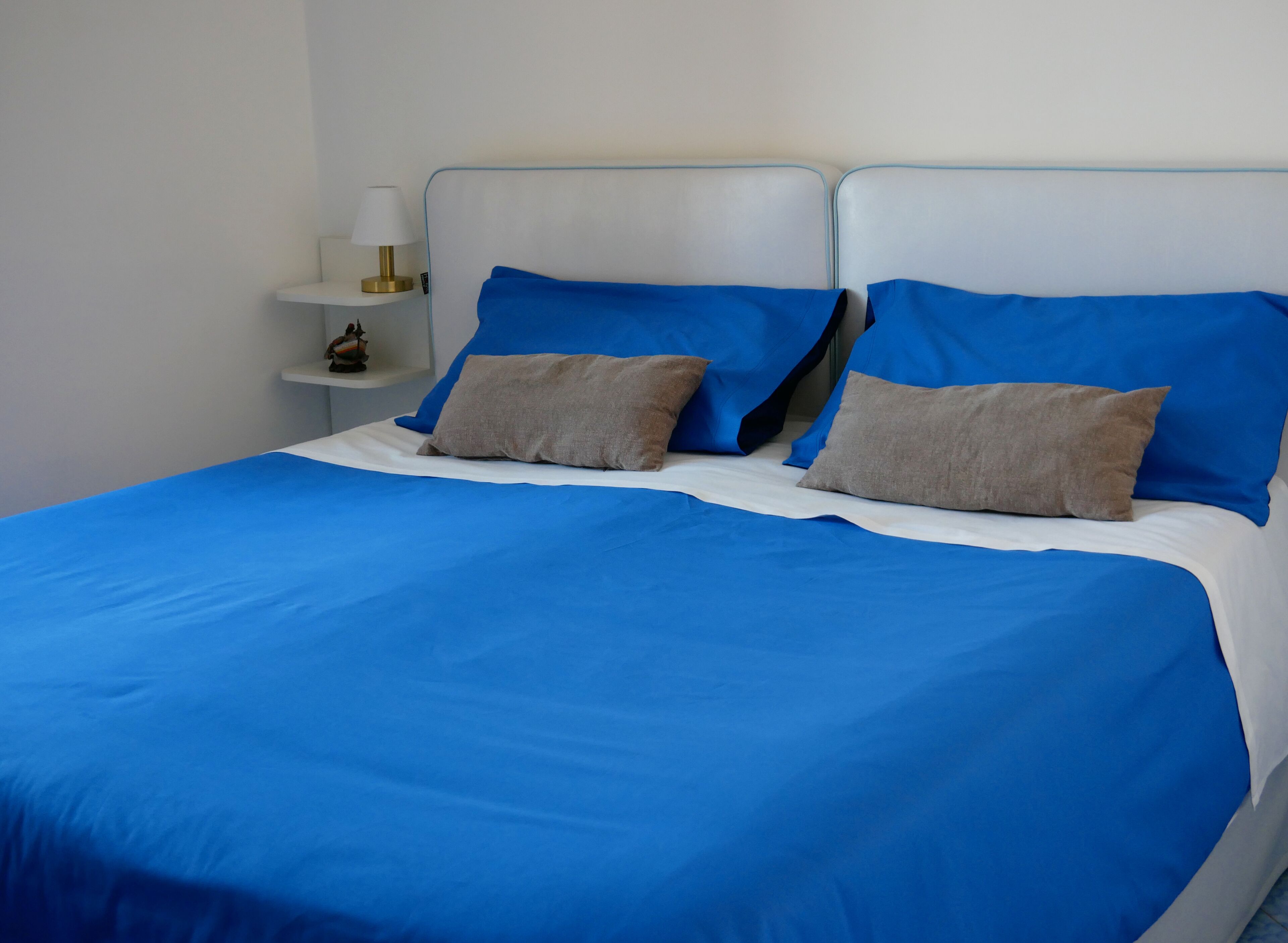Ein Bett mit blauer Bettwäsche und zwei Kissen, ein Nachttisch mit Lampe und ein kleines Regal mit einem dekorativen Gegenstand.