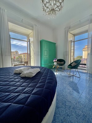 Un dormitorio con una cama grande, un armario verde, dos sillas y vistas a la ciudad a través de ventanas abiertas.