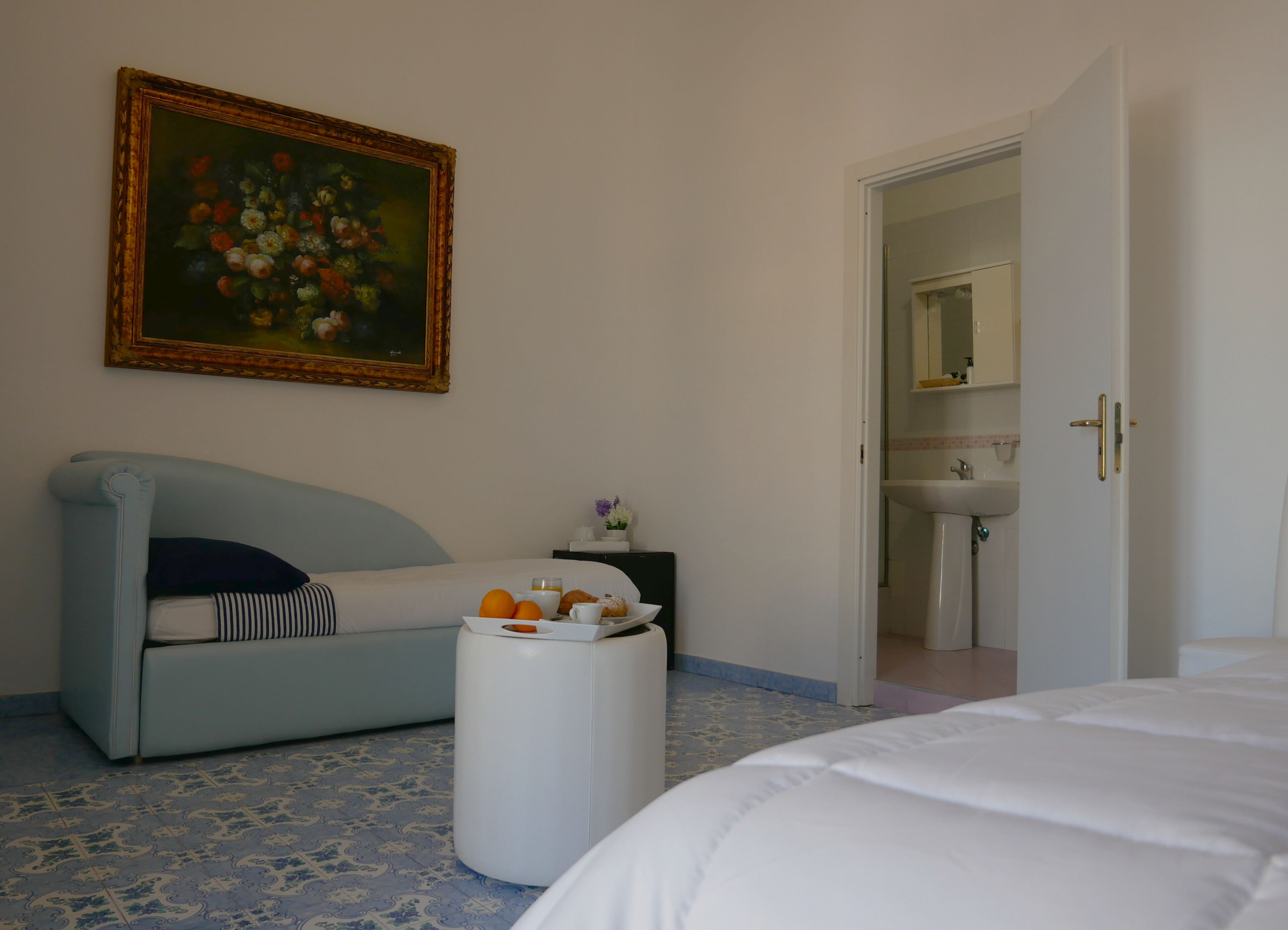 Ein Hotelzimmer mit einem hellblauen Sofa, einem Nachttisch mit einer Schale Orangen und Gebäck, einem Bild an der Wand und einem durch eine geöffnete Tür einsehbaren Badezimmer.