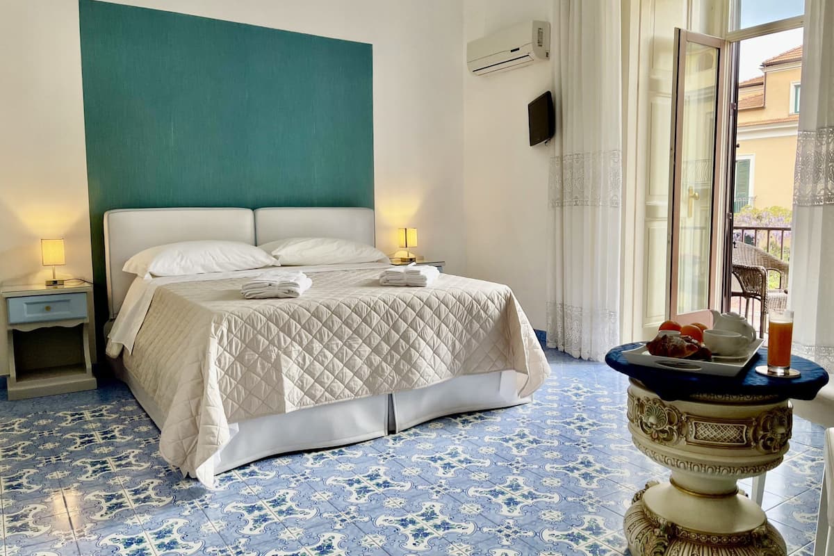 Una habitación de hotel con una cama, lámparas de noche, una mesita decorativa con una bebida y baldosas de suelo estampadas.