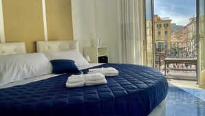 Ein großes Bett mit dunkelblauer Decke, weißen Kissen und einem weißen Kopfteil. Ein kleiner Nachttisch mit Lampe und Steckdose steht daneben. Das Zimmer hat ein großes Fenster mit weißen Vorhängen und bietet einen Blick auf Gebäude und eine Straße.